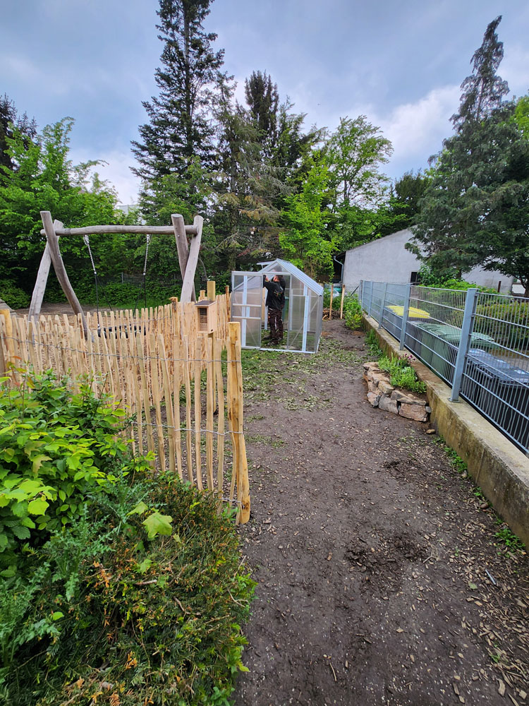 Schulgarten Meckenheim: fertiger Garten mit Vitavia Gewächshaus Planet, Modell 5000  in Alu und Hohlkammerplatten umgeben von Holzzaun